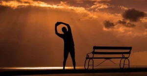 A man stretching in front of a bench, in a dark sunset setting.