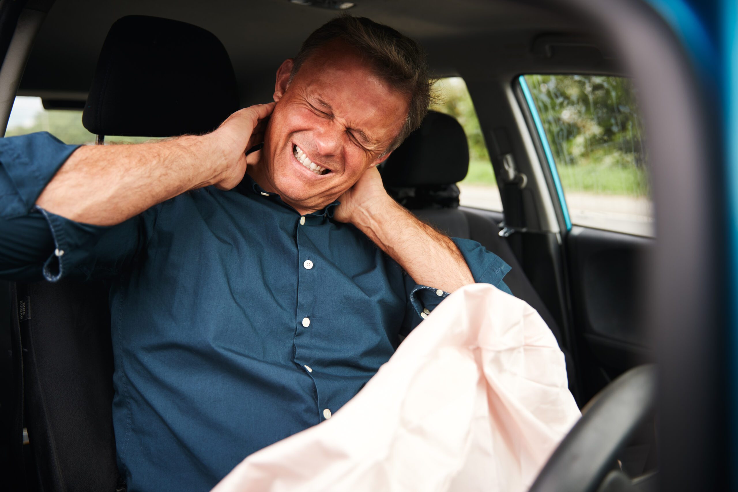 A man sitting in a car, touching his neck with both hands and looking to be in great pain.