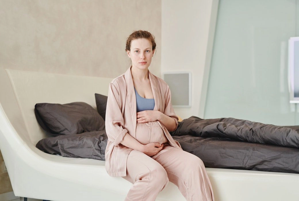 A pregnant woman sitting on a bed and holding her belly while looking at the camera.
