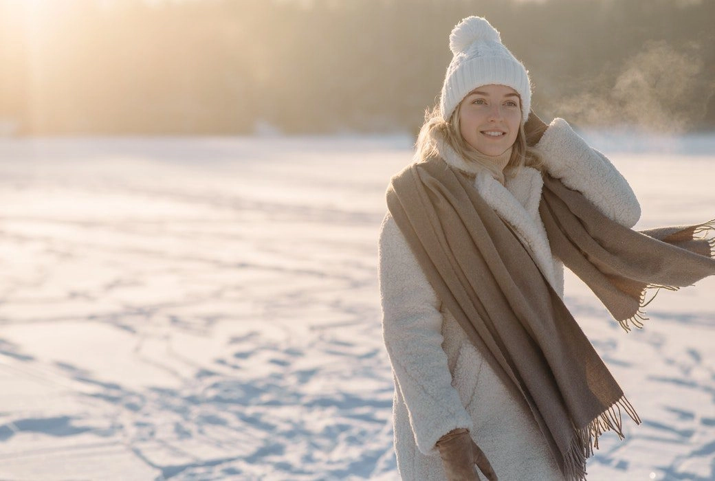 A woman standing in a winter outdoor scenery, wearing white and beige winter clothing and looking happy/