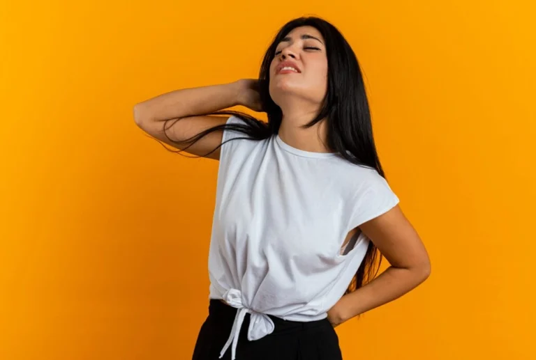 A woman standing in front of an orange background, holding her neck with one hand and her lower back with the other, looking to be in pain.