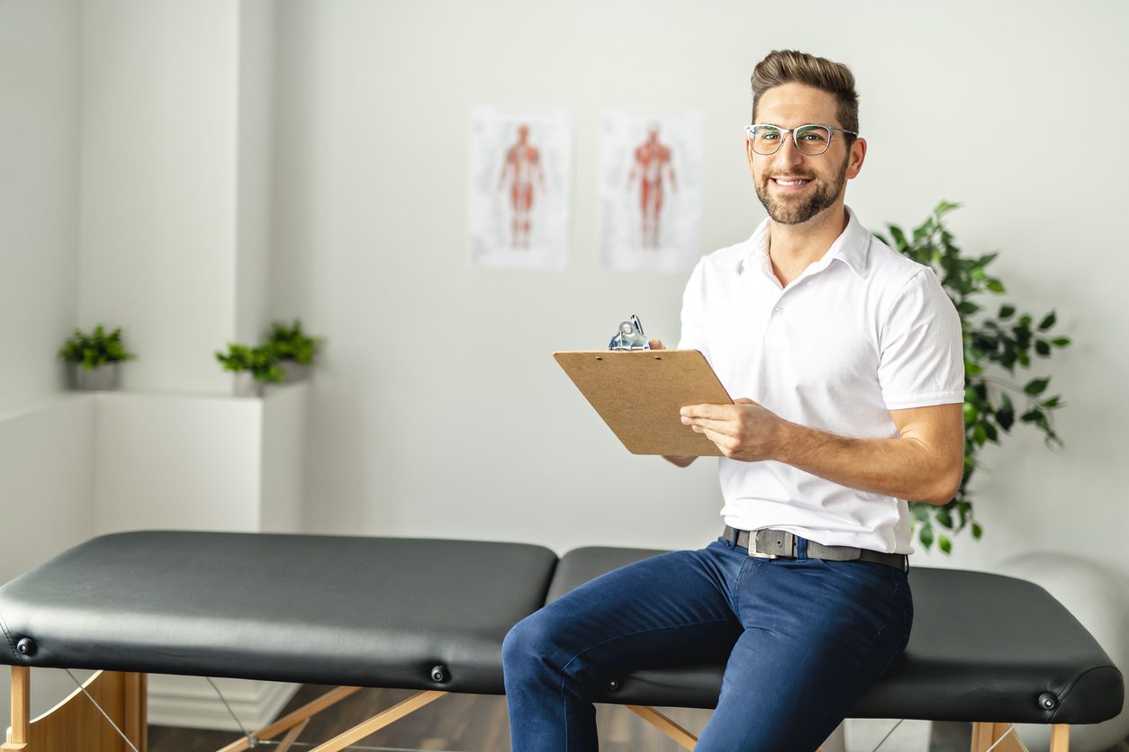 A happy chiropractor sitting on a massage table holding a notepad.