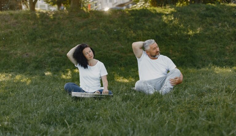 Two people doing neck stretches while sitting down on an outdoor scenery.