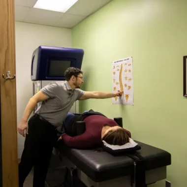 A chiropractor explaining a diagram of the spine to a patient lying in a treatment room.