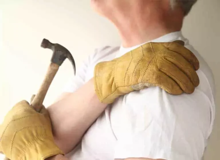 A man with working gloves holding a hammer and touching his shoulder.