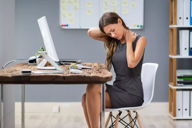 A woman having pain in her neck sitting in front of a desk with a computer.