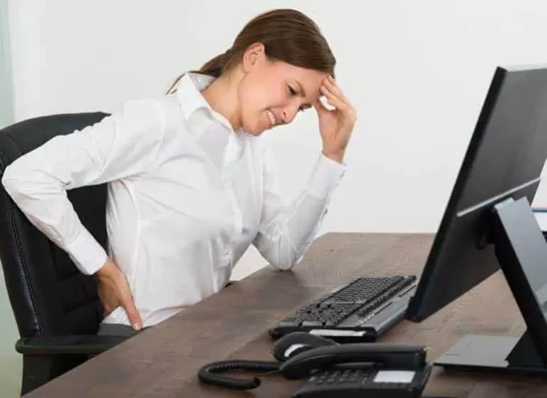 A woman touching her head and back in pain in front of a computer.